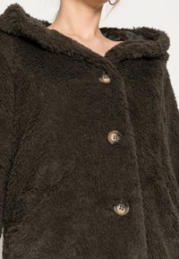 Brun pelsforet frakke med hætte, der har store knapper, blød tekstur og en løs pasform. Bemærkelsesværdige varme- og komfortelementer.