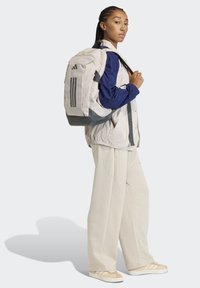 Mochila de color gris claro con detalles en gris oscuro y tres franjas negras. El modelo lleva una camisa de manga larga azul y pantalones color crema, mostrando un estilo casual.