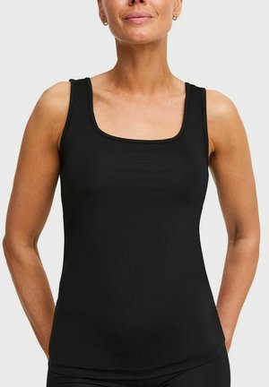 Vrouw draagt een strakke zwarte mouwloze tanktop met een vierkante halslijn, staand tegen een effen lichte achtergrond.