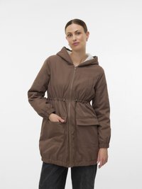 Woman wearing a brown hooded jacket with front zipper and pockets, standing with one hand in pocket against a plain white background.