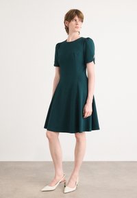 Teal dress with short puff sleeves, fitted bodice, and flared skirt. Made from a structured fabric. White pointed-toe slingback shoes.