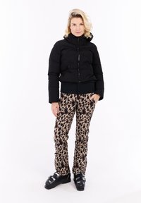 Mujer con chaqueta de plumas negra, pantalones de esquí con estampado de leopardo y botas de esquí negras, de pie contra un fondo blanco.