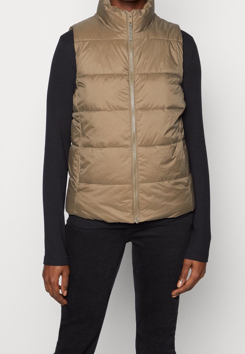 Helles Taupe ärmelloses Puffer-Weste mit vertikalen Nähte, hohem Kragen und einem Front-Reißverschluss, kombiniert mit einem schwarzen Langarmoberteil.
