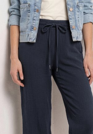 Mujer con chaqueta vaquera clara y pantalones azul marino con cordón, de pie frente a una pared blanca texturizada.