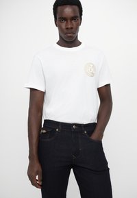 Λευκή βαμβακερή μπλούζα με στρογγυλή λαιμόκοψη και χρυσό έμβλημα, σε συνδυασμό με σκούρα τζιν παντελόνια με χρυσές ραφές και μπροστινές τσέπες.