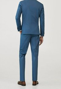 Uomo che indossa un completo blu aderente con pantaloni abbinati e scarpe eleganti marroni, in piedi con le mani rilassate ai lati, vista posteriore.