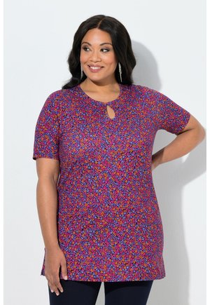 Frau trägt eine violette Tunika mit kurzen Ärmeln und floralem Muster, einem kleinen Schlitz am Ausschnitt, lächelt und blickt leicht seitlich.