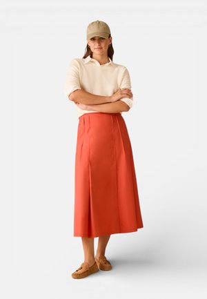 Donna che indossa un cappellino beige, una maglia a maniche lunghe color crema, una gonna midi arancione e mocassini marroni, in piedi con le braccia incrociate.