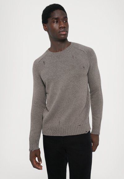 Pull en maille grise avec un col rond, des poignets et un ourlet côtelés, présentant de petits détails usés sur l'ensemble du tissu. Associé à un pantalon sombre.