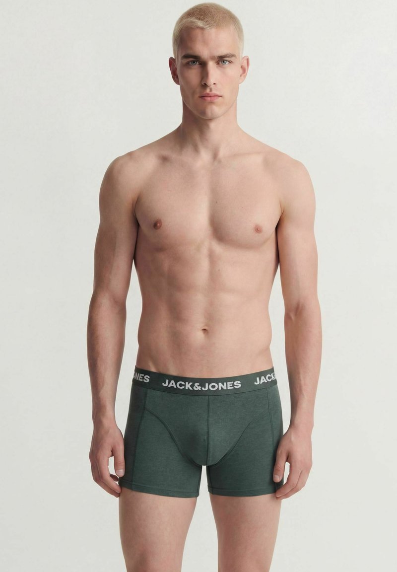 Jeune homme aux cheveux blonds courts portant un boxer vert foncé Jack & Jones, debout devant un fond clair uni.