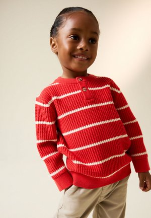 Junges Kind lächelt und trägt einen roten Strickpullover mit weißen Streifen und beige Hose vor einem schlichten hellen Hintergrund.