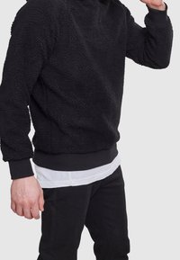 Persona che indossa un maglione in pile nero testurizzato sopra una camicia bianca con pantaloni neri, in posa contro uno sfondo grigio chiaro uniforme.