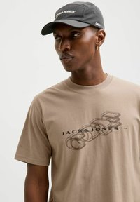 Beżowy bawełnowy t-shirt z grafiką przedstawiającą kontury i tekst "JACK & JONES". Noszony z czarną czapką, na której widnieje nazwa marki.