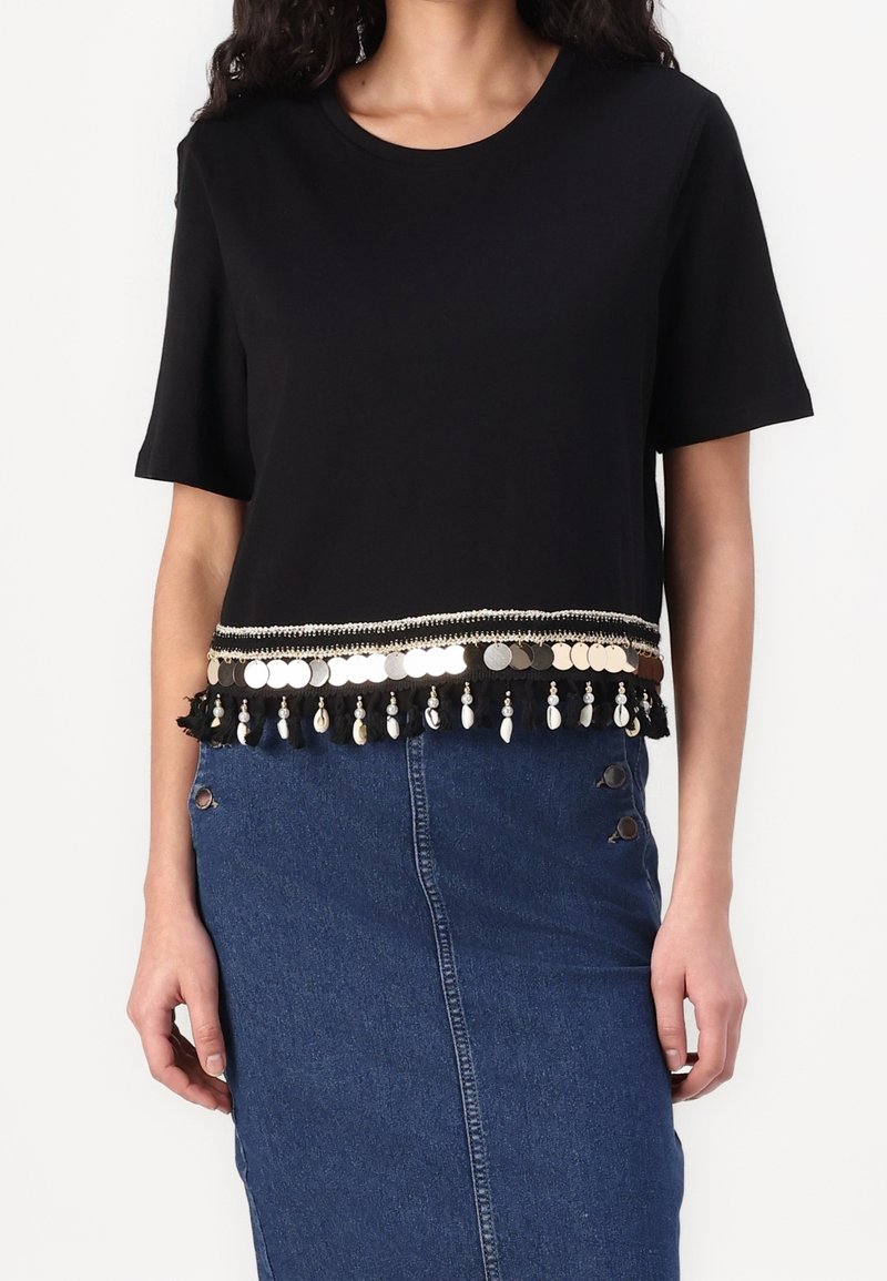 Camiseta corta negra de manga corta, con un borde decorativo de flecos con discos metálicos y borlas en el dobladillo. Combinada con una falda de mezclilla.