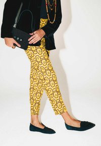 Blazer in velluto nero, pantaloni gialli con motivo a curve marroni, ballerine azzurre impreziosite da perline. La modella tiene in mano una pochette nera con perline.