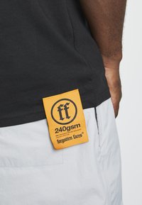 Svart t-skjorte med tekstur, med et gult merket som viser "240gsm" og "forgotten faces"-logoen. Merket er sydd på siden.