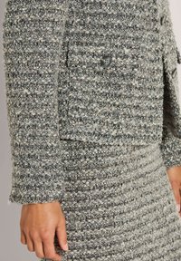 Grau-weiße, strukturierte Tweed-Jacke und -Rock mit Knopfdetails, getragen von einer Person mit entspannten Händen an den Seiten.
