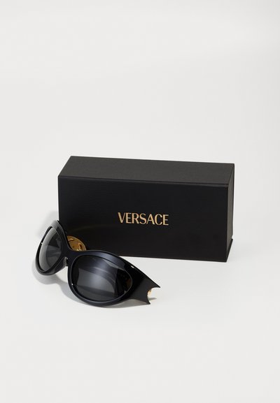 Occhiali da sole Versace cat-eye neri con lenti scure accanto a una scatola nera con il logo Versace dorato su sfondo bianco.