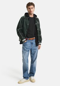 Grøn baseballjakke med hvid piping, sort hættetrøje under, sort t-shirt, lyseblå jeans og hvide sneakers med brune accenter.