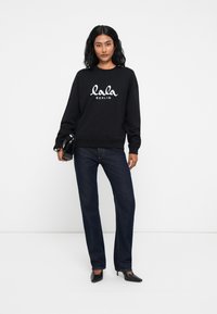 Čierna mikina s bielym logom "lala BERLIN", v kombinácii s tmavými džínsami. Model drží malú čiernu kabelku; celok dopĺňajú lodičky.