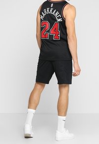Maillot de basket-ball noir avec le nom "MARKKANEN" et le numéro "24" en lettres rouges, associé à un short noir et des baskets blanches, posé devant un fond uni.