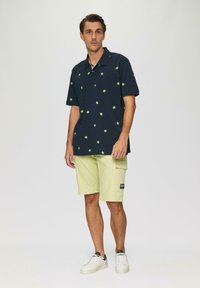 Granatowa koszulka polo w zielony wzór palm, zestawiona z jasnobeżowymi szortami cargo i białymi trampkami z czarnymi akcentami.