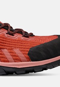 Chaussure de sport rouge avec une tige en maille texturée, un embout en caoutchouc noir, une semelle intermédiaire épaisse rose, et des lacets noirs contrastants avec des accents orange.