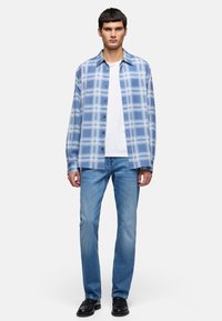 Homme debout portant une chemise à carreaux bleu clair à boutons par-dessus un t-shirt blanc, un jean bleu et des chaussures noires sur un fond uni.