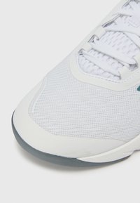 Hvid sportssko med en struktureret mesh-overdel, gummisål og flade snørebånd. Har en polstret krave og minimale dekorative detaljer.