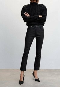 Černý pletený svetr s vysokým límcem a texturovaným vzorem, spárovaný s úzkými černými džínami a špičatými černými lodičkami.