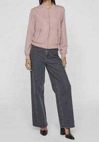 Femme portant une veste zippée rose clair, un jean large gris foncé et des chaussures à talons noires, debout devant un fond uni.