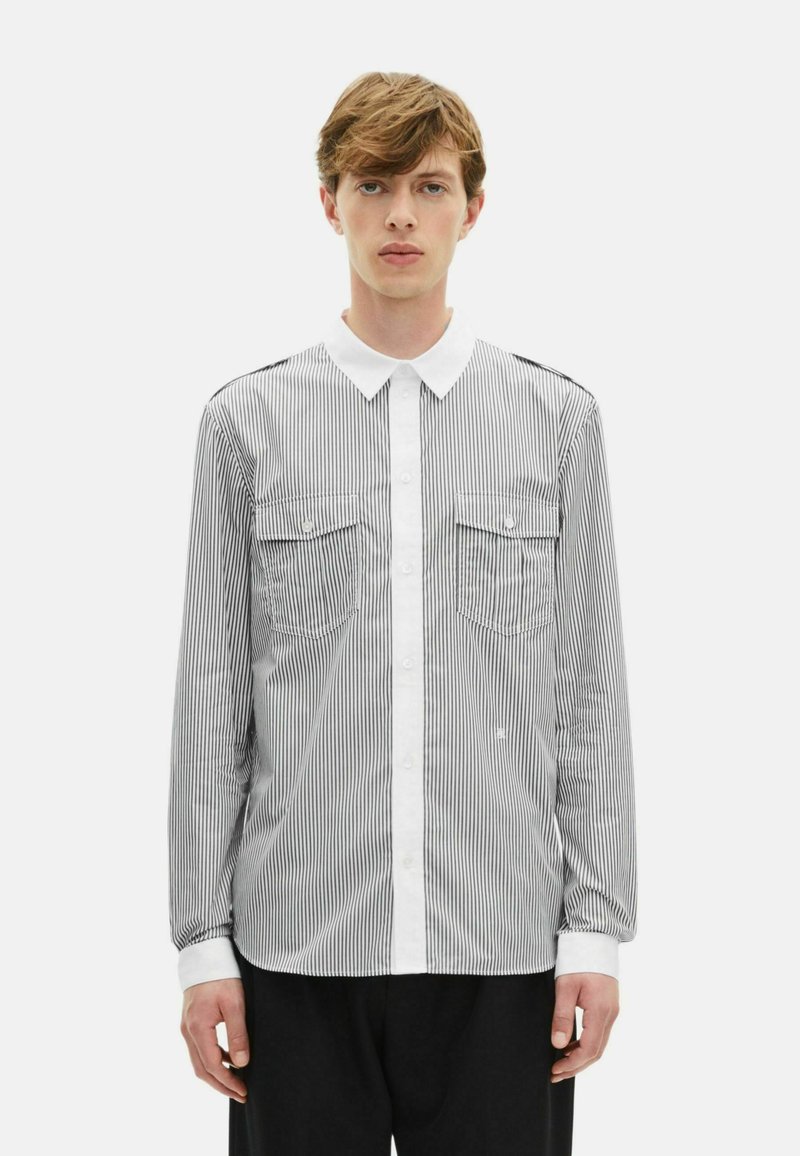 Ριγέ πουκάμισο μακρυμάνικο με λευκό γιακά που κουμπώνει, δύο τσέπες στο στήθος και αντίθετους λευκούς καρπούς, με μαύρες και λευκές κατακόρυφες ρίγες.