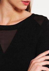 Svart tröja med en genomskinlig v-ringning, texturerad tyg och en subtil glans längs halslinningen. Närbild som visar axeln och handen.