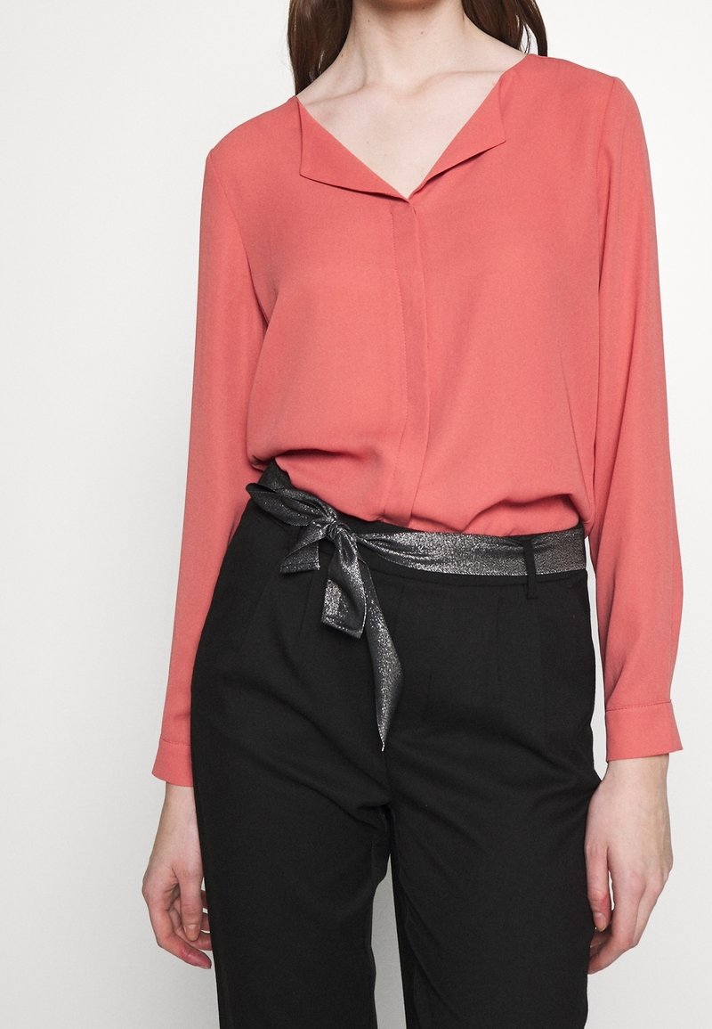 Femme portant un chemisier corail à manches longues avec un col et un pantalon noir attaché avec une ceinture en tissu noir brillant nouée en nœud.