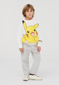 Felpa bianca con grande grafica di Pikachu gialla, pantaloni della tuta grigi e sneakers bianche. Presenta polsini e orlo a costine. Design semplice.