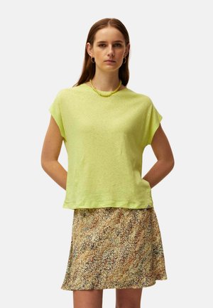 Mujer con cabello liso y castaño lleva una blusa suelta de manga corta color verde lima, collar de cuentas y falda estampada hasta la rodilla con bolsillos.