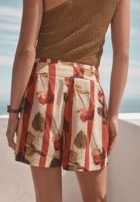 Shorts à motif floral en crème, rouge et vert, avec des plis latéraux et une taille haute. Confectionnés en tissu léger.