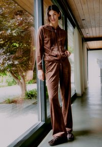 Ensemble de survêtement à capuche en velours brun avec fermeture éclair, doté d'une poche avant. Pantalons ajustés avec une coupe évasée. Texture lisse, devant un mur en verre.