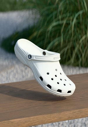 Hvid plastiktræsko med ventilationshuller, rundet design og tekstureret sål. Crocs-logo på remmen. Placeret over en træoverflade.