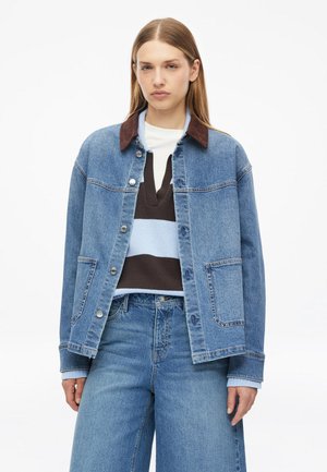 Junge Frau mit glattem blondem Haar, die eine blaue Jeansjacke, einen gestreiften Pullover und hoch taillierte Jeans vor einem einfarbigen Hintergrund trägt.