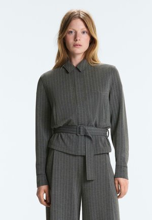 Rdečelasa ženska, oblečena v sivi jopič s črtami in pasom ter ujemajoče hlače, stoji pred enostavnim svetlim ozadjem.