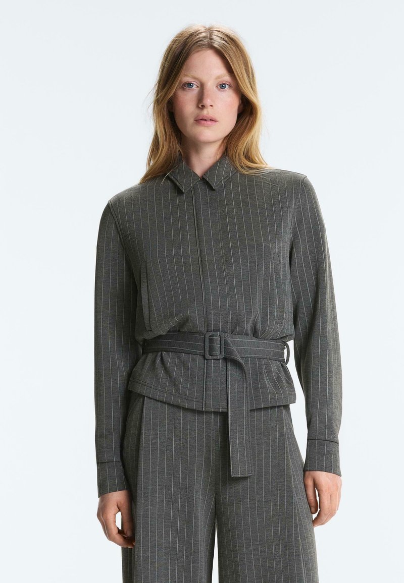 Femme aux cheveux roux portant une veste grise à fines rayures avec ceinture et un pantalon assorti, debout devant un fond clair uni.