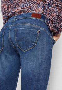 Modré džíny z denimu s hladkým povrchem, s koženou nášivkou v pase a dvěma zadními kapsami s dekorativním prošitím.