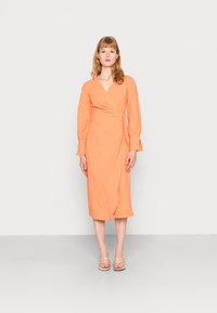 Vestido envelopado em tecido laranja brilhante com uma superfície texturizada, mangas longas e um detalhe de laço na cintura, terminando na meia panturrilha.