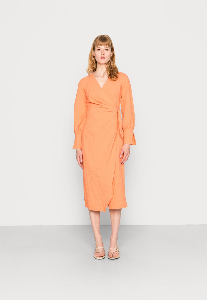 Vestido envelopado em tecido laranja brilhante com uma superfície texturizada, mangas longas e um detalhe de laço na cintura, terminando na meia panturrilha.