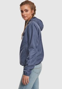 Könnyű kék kapucnis dzseki, rugalmas mandzsettákkal és laza szabással. Simább, vízlepergető anyagból készült, a kényelem érdekében.