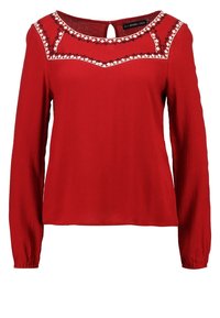Rode blouse met lange mouwen en een ronde hals. Voorzien van decoratieve witte stiksels langs de schoudernaad. Gemaakt van zachte stof met een ontspannen pasvorm.