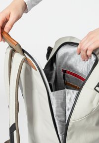 Zaino grigio con dettagli in pelle marrone; l'interno presenta una fodera a scacchi e una tasca con cerniera rossa; progettato per la praticità e l'organizzazione.