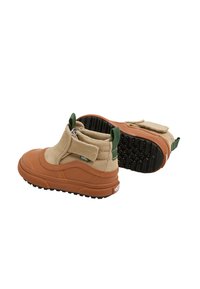 Un par de botines Vans en tonos marrones y beige con tiradores verdes, suelas de goma negra y correas de velcro sobre un fondo blanco.