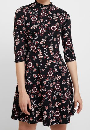 Robe noire avec motif floral rouge et blanc, taille ajustée, manches trois-quarts, et un petit col à volants sur une personne debout.